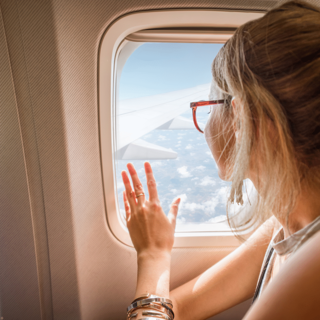 Woman smiling and waving at the airplane window mid-flight, with clouds and wing visible outside.