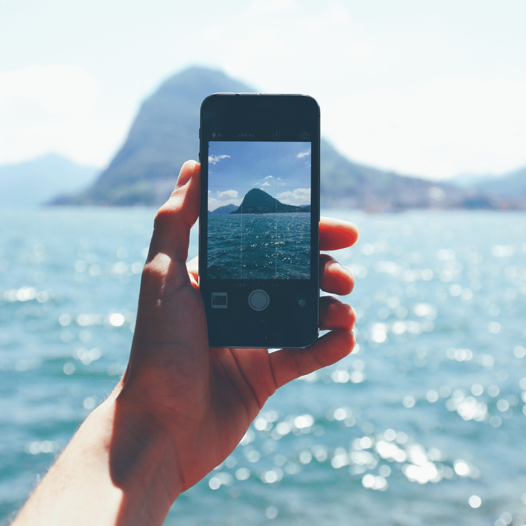 Hand holding a smartphone taking a photo of a mountain across the ocean on a sunny day.