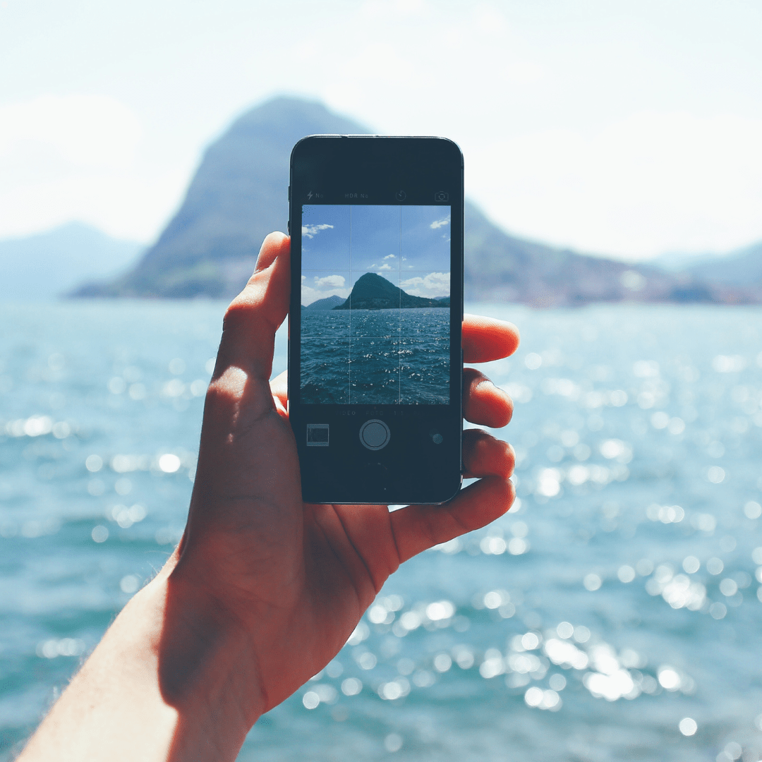 Hand holding a smartphone taking a photo of a mountain across the ocean on a sunny day.
