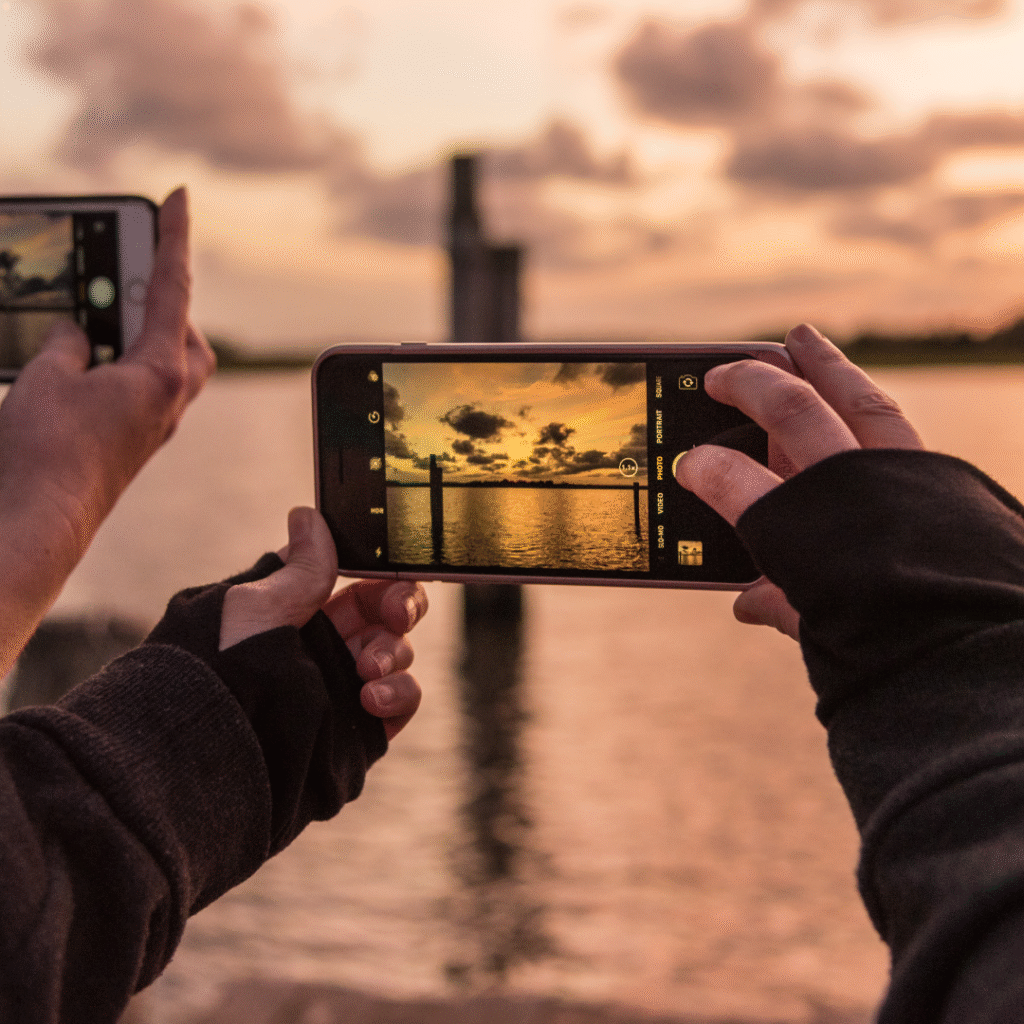 Hands holding a smartphone to photograph a sunset over water, symbolizing storytelling and reflection.