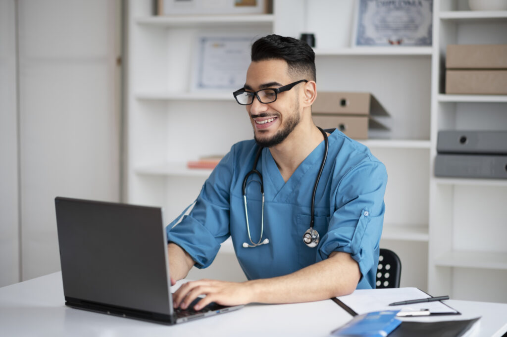 Medical professional in scrubs smiling while working on a laptop in an office.