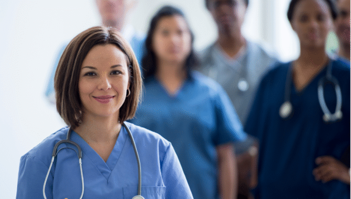 A smiling bilingual nurse standing confidently in front of a diverse medical team, representing the importance of communication and cultural connection in Canadian healthcare.