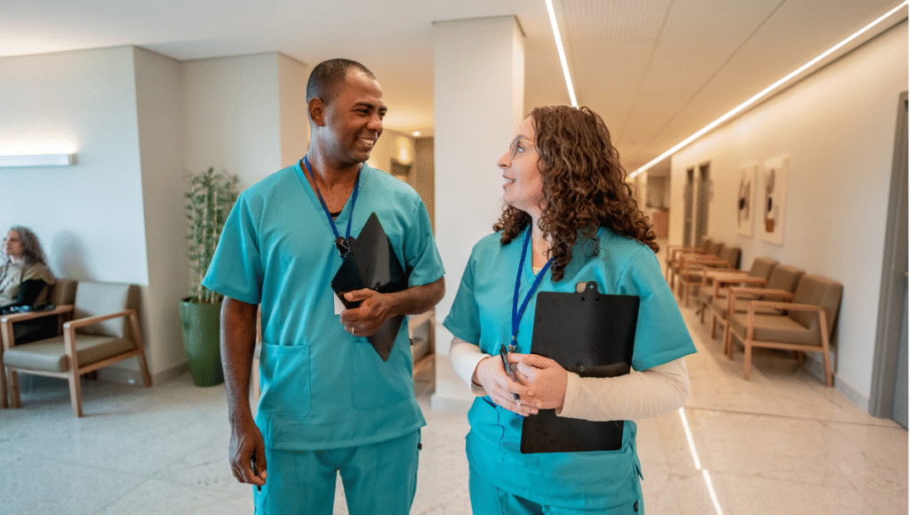 Two travel nurses in scrubs walking and talking in a hospital hallway, discussing assignments and patient care
